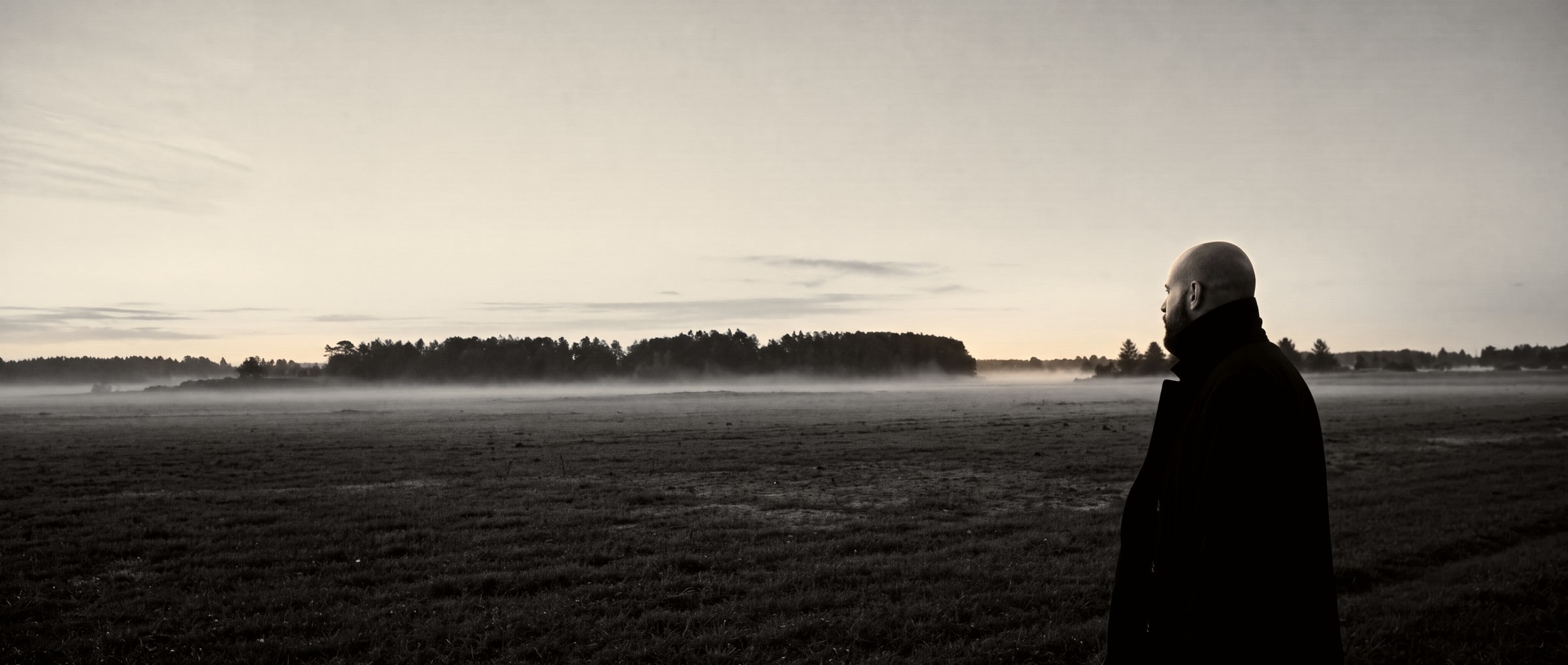 A misty field at dawn, a figure in a long coat at its edge.