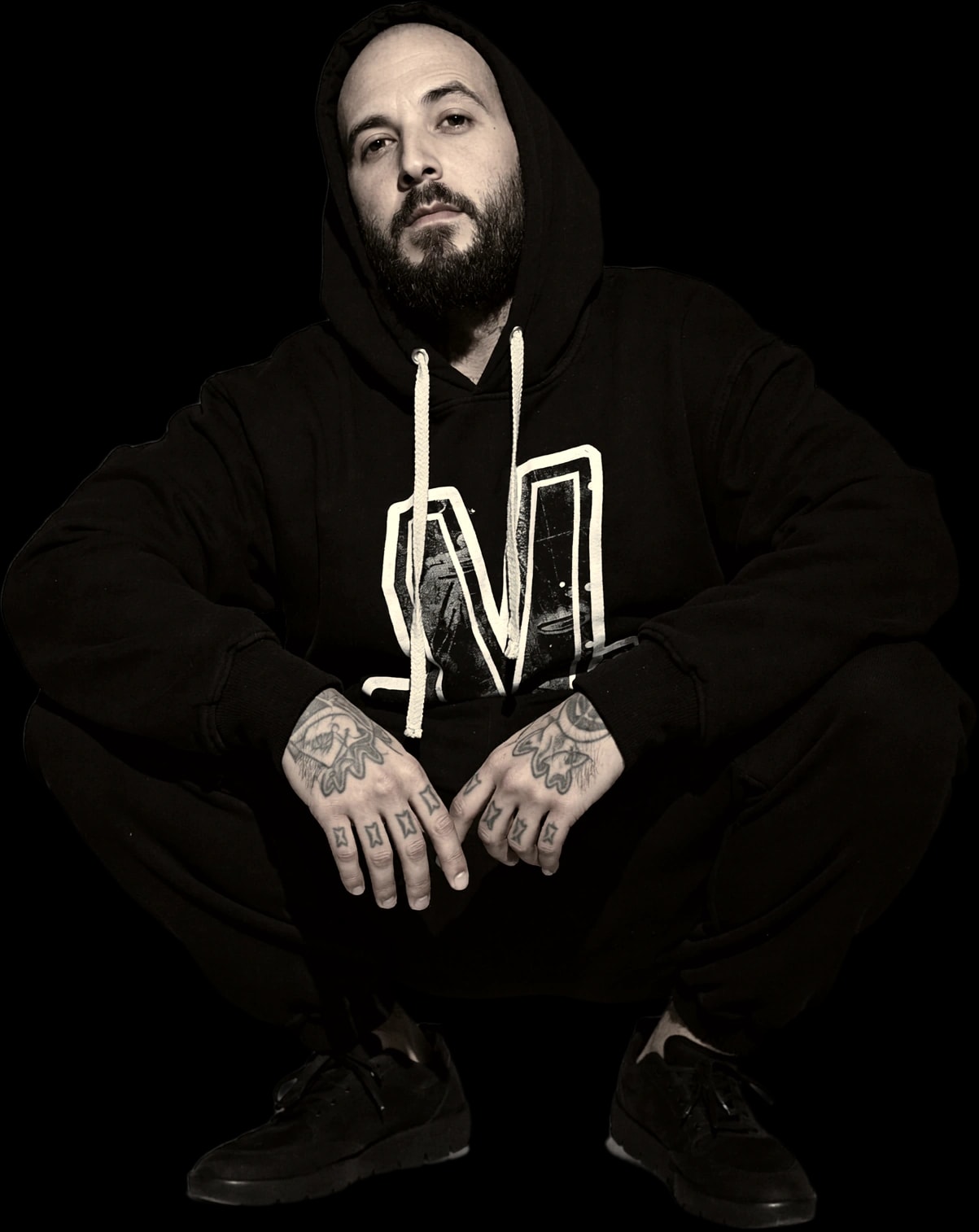 Nicolás Morin crouching in a black hoodie, his tattooed hands resting together in front of him.
