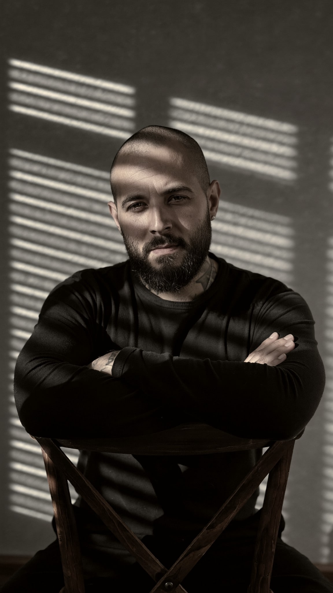 Nicolás Morin seated backwards on a wooden chair, arms crossed, blind shadows on the wall behind him.