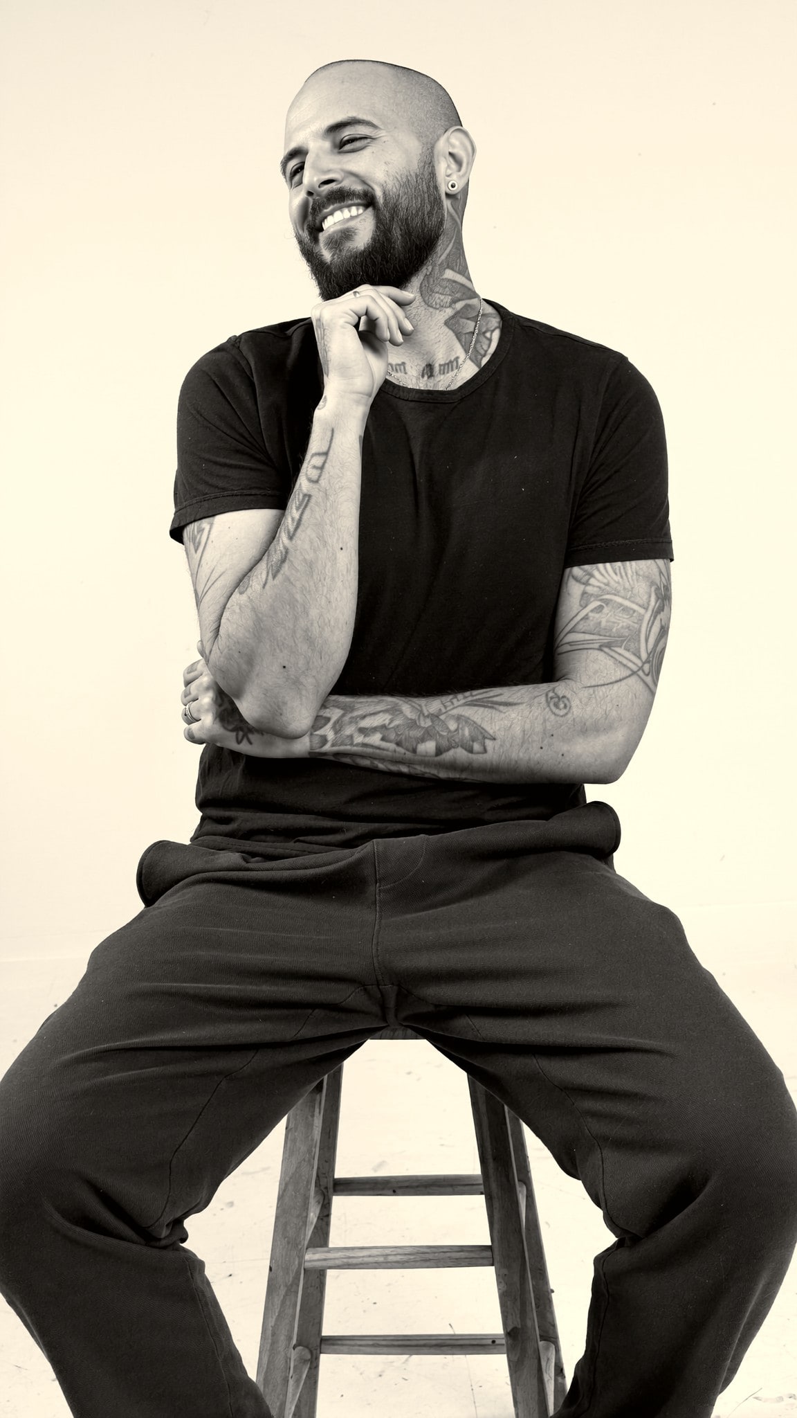 Nicolás Morin laughing on a wooden stool, his arms and neck tattooed.