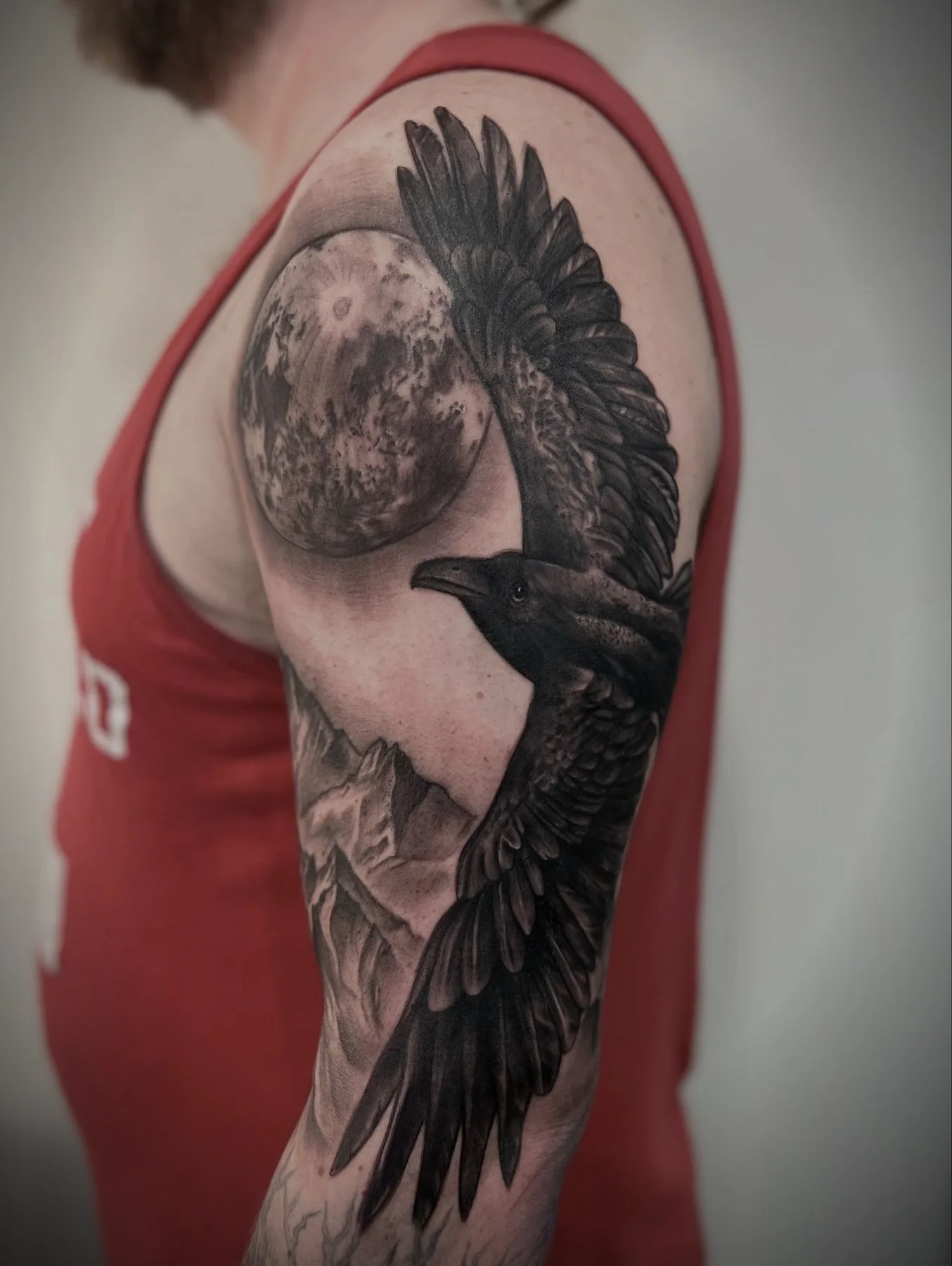 A raven in flight beneath a full moon, tattooed across an upper arm in black and grey realism.