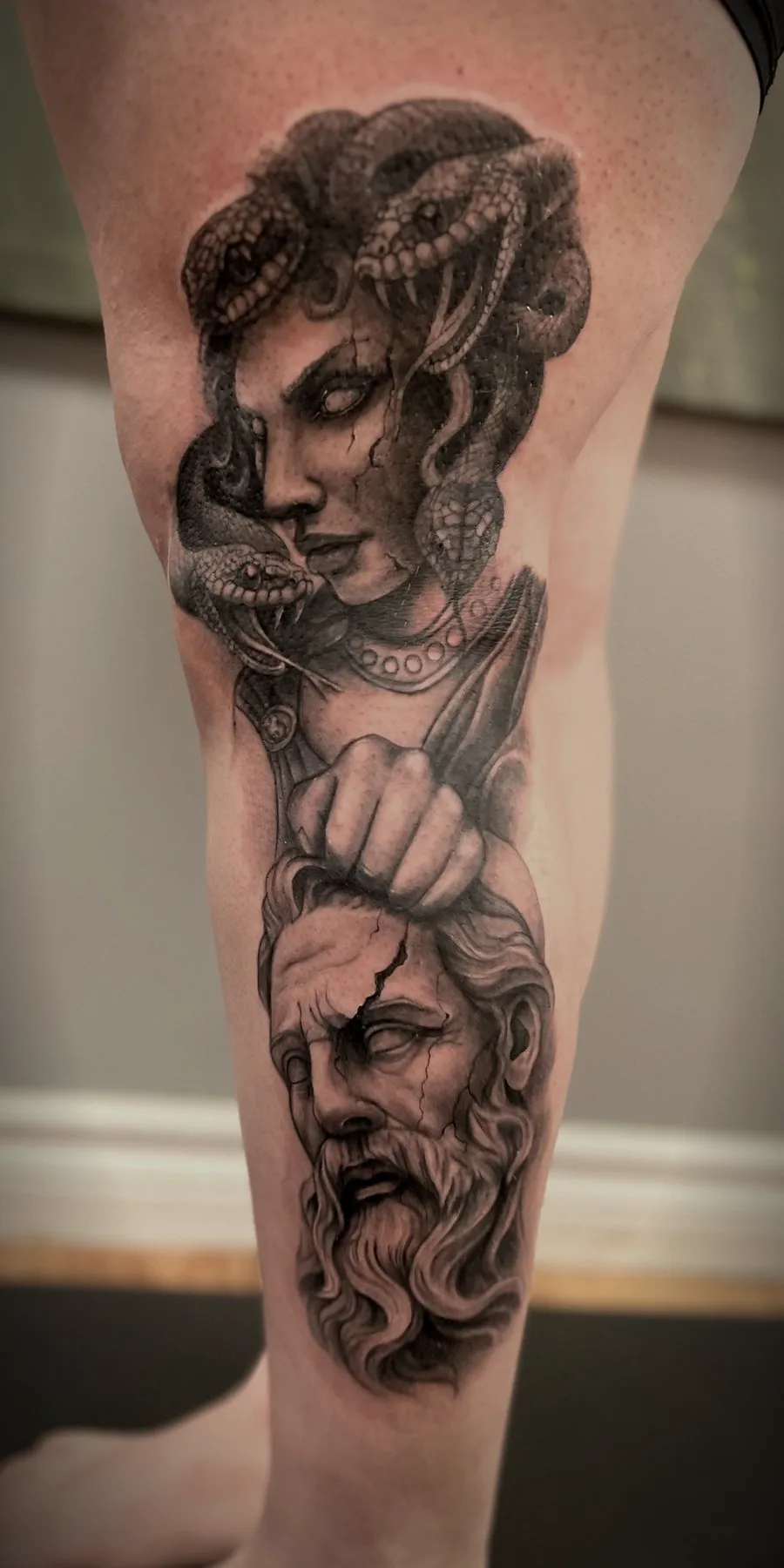 A calf tattooed in black and grey realism: Medusa with snakes coiled through her hair, holding the severed stone head of a bearded god.
