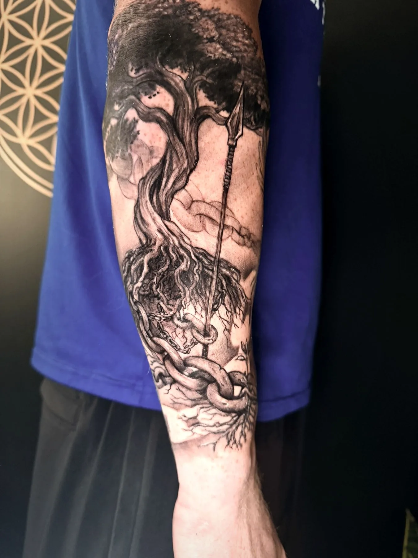 A Norse warrior beneath a tree and a spear, tattooed along a forearm in black and grey realism.