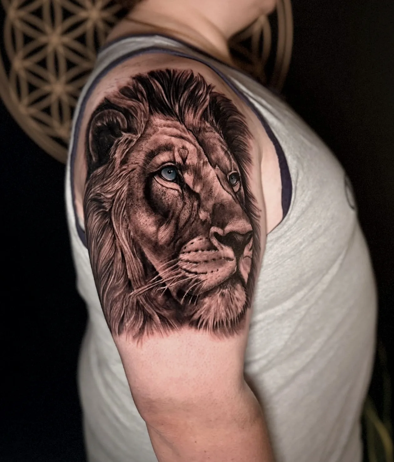 A lion's head tattooed on an upper arm in black and grey realism, its mane rendered hair by hair, one eye left pale blue.