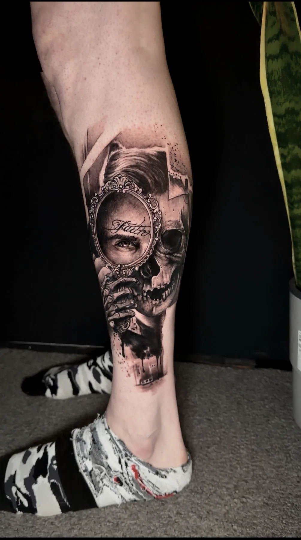 A skull reflected in a hand mirror, tattooed down a shin in black and grey realism.
