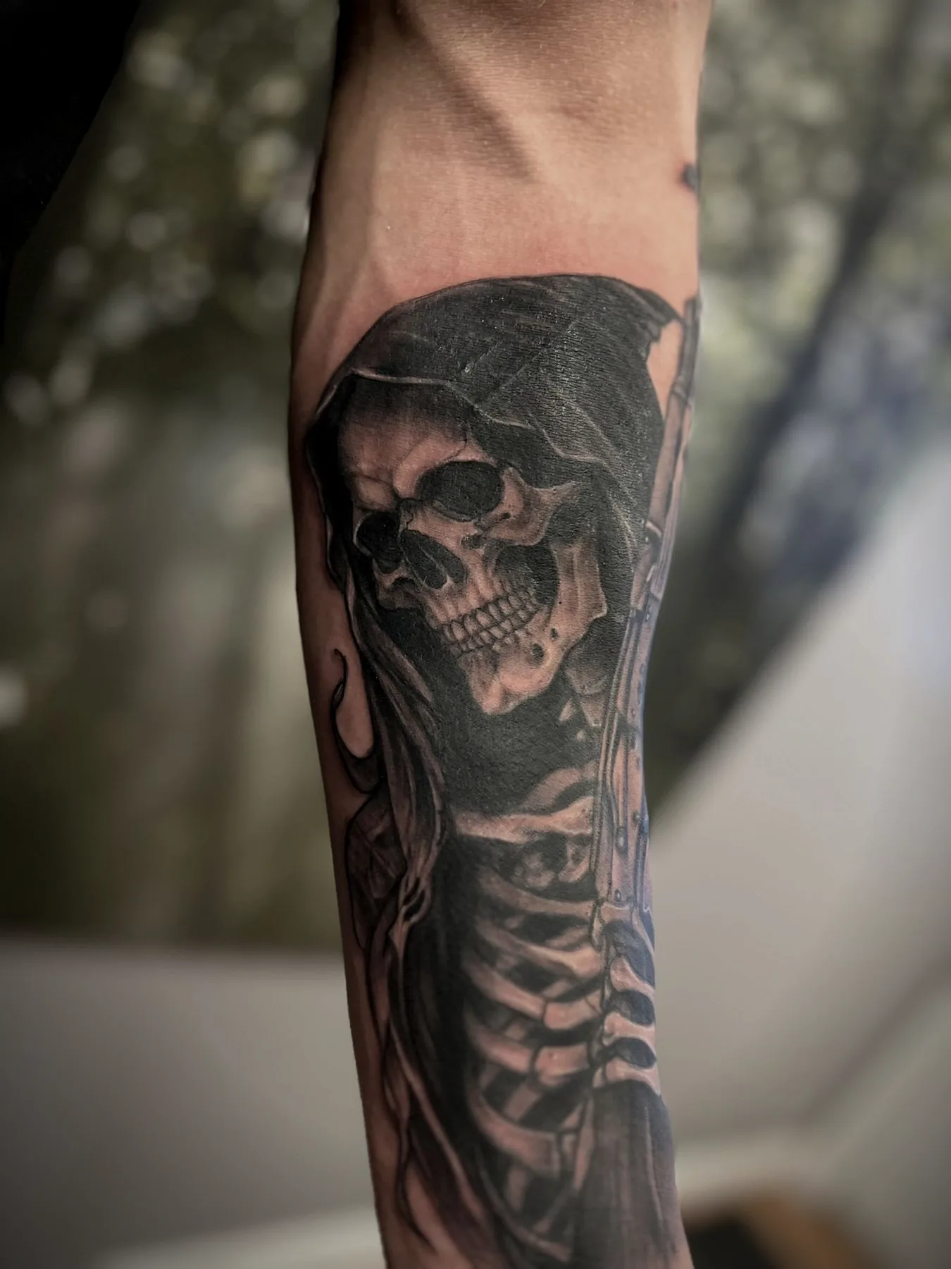 A hooded reaper over a skeletal ribcage, tattooed along an arm in black and grey.