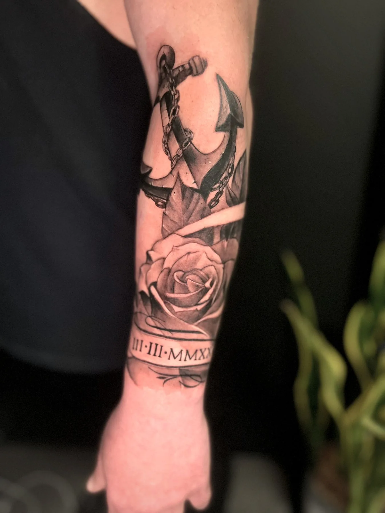 A rose pierced by a dagger and an anchor, tattooed along a forearm above a date in Roman numerals.