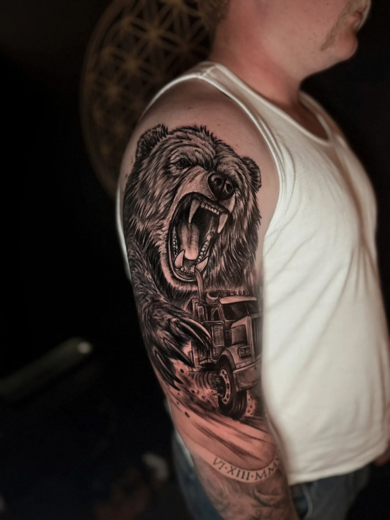 A roaring grizzly above a semi-truck charging out of a dust cloud, with a Roman-numeral date beneath, tattooed on an upper arm in black and grey realism.