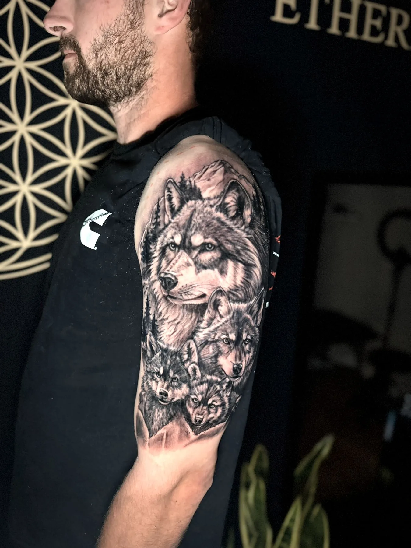 A wolf with her cubs gathered beneath her, set against mountains and pines, tattooed on an upper arm in black and grey realism.