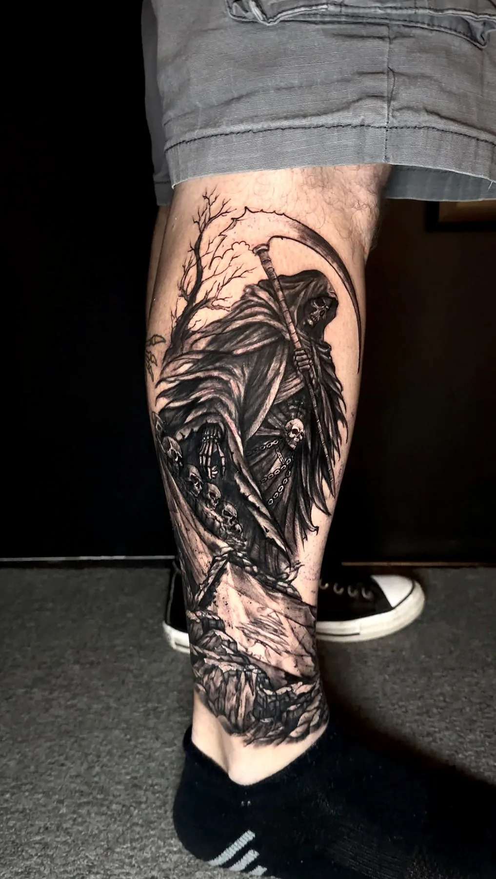 A hooded reaper with his scythe, walking past a bare tree and a trail of skulls, tattooed down a calf in black and grey.