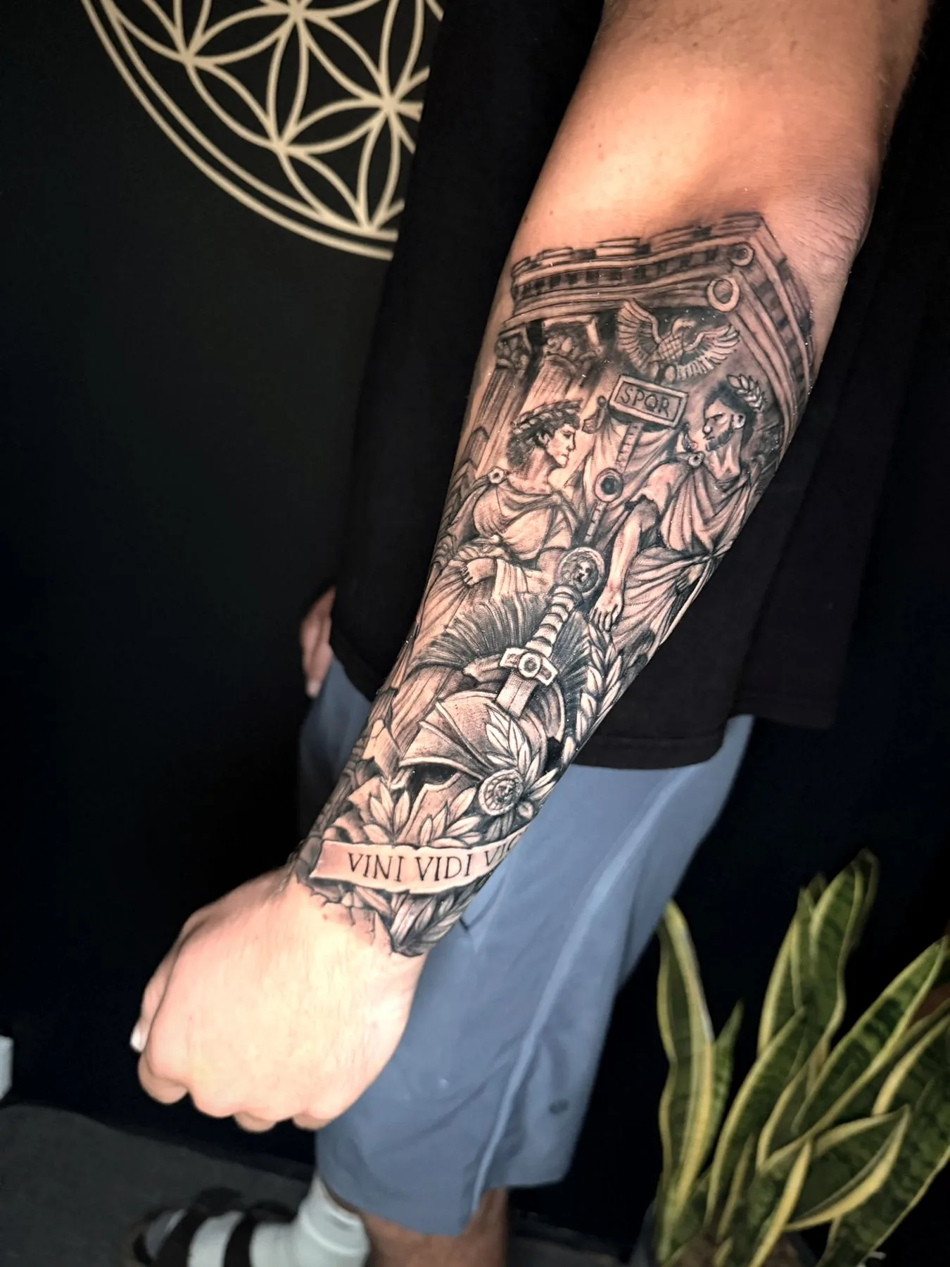 A Roman scene of a temple, an SPQR standard, a helmet and a sword over a laurel banner, tattooed on a forearm in black and grey realism.