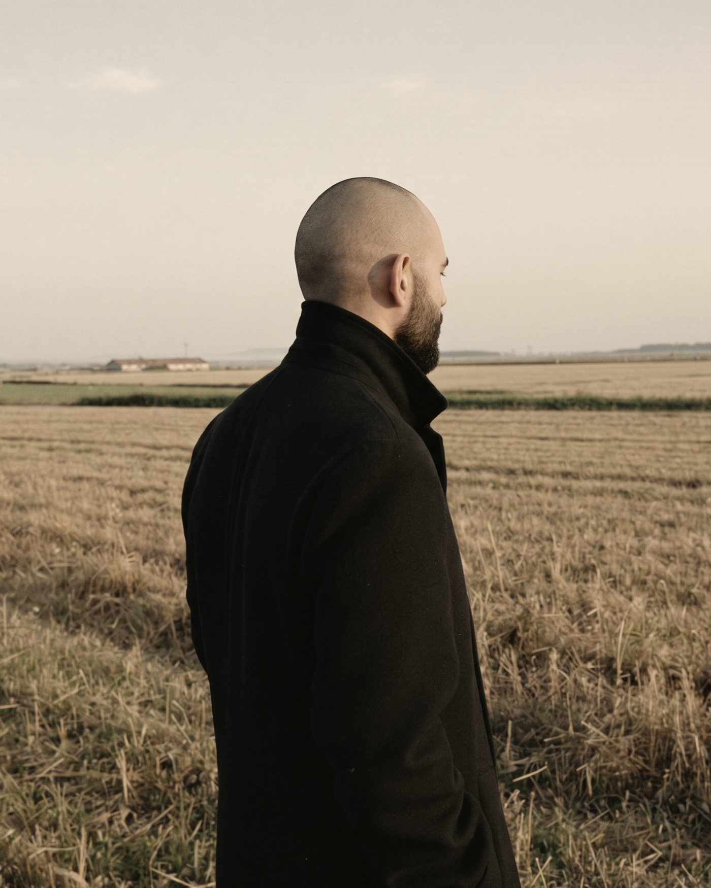 A generated image of Nicolás Morin in a long black coat, standing at the edge of a harvested field in warm evening light, looking away.