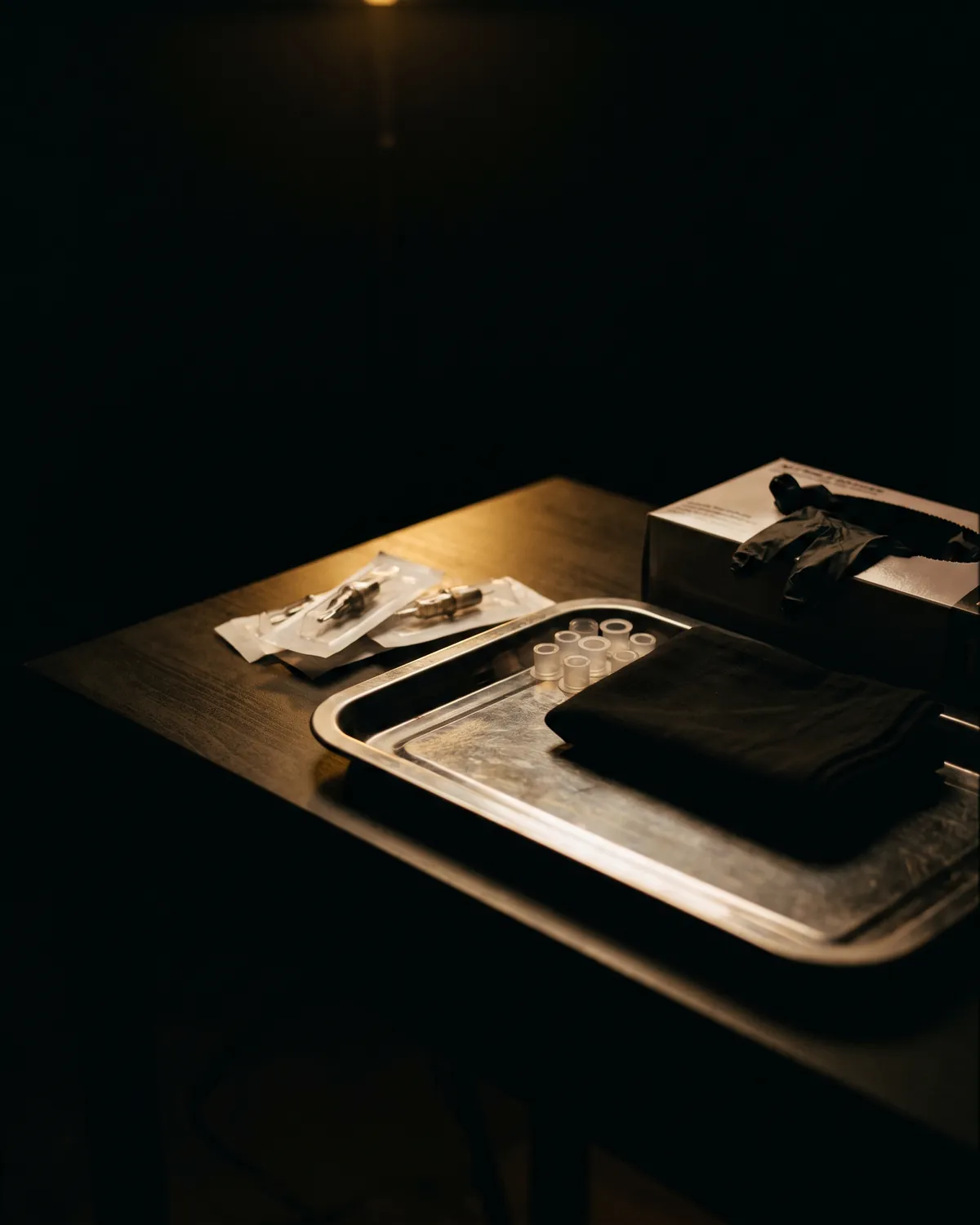 A steel tray on a dark workbench under one warm lamp, laid out with sealed needle cartridges, empty ink caps, a folded black cloth and a box of gloves.